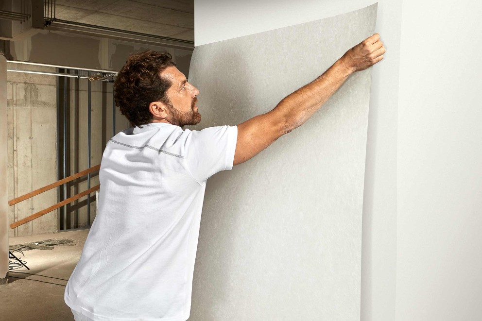 Mann befestigt Malervlies an einer Wand in einem Raum