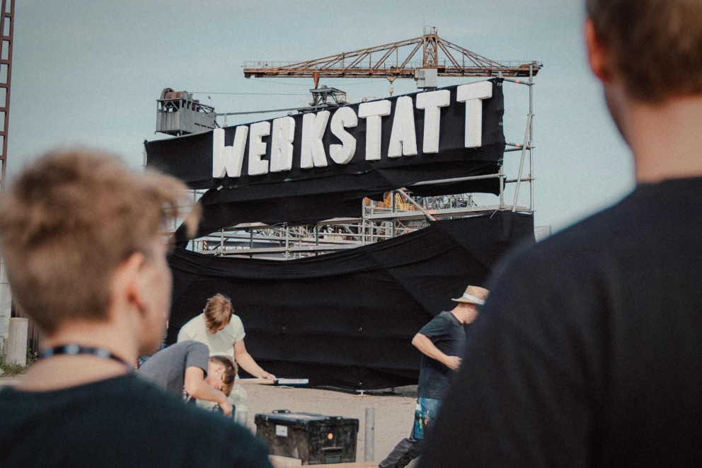 Szene mit der Aufschrift Werkstatt und einer Krananlage im Hintergrund.