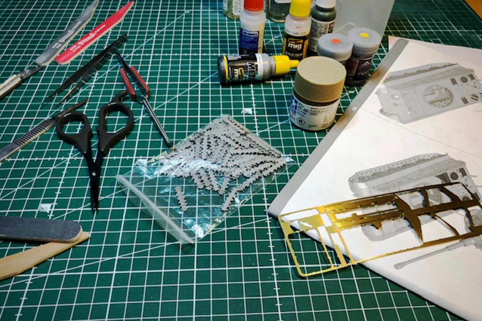 Modellbau-Werkzeuge und Zubehör auf einer Schneidematte, einschließlich Messer, Scheren, Pinzetten, Feilen, Klebstoff, Farbe, einem Beutel mit Kettenmodellbauteilen, einer Platine mit Metallmodellbauteilen und einer Bauanleitung