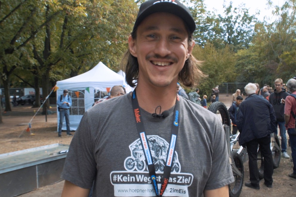Porträt eines lächelnden Mannes mit Bart und Kappe, der ein T-Shirt mit dem Aufdruck '#KeinWegItAsZiel' und ein Lanyard der Maker Faire Hannover trägt