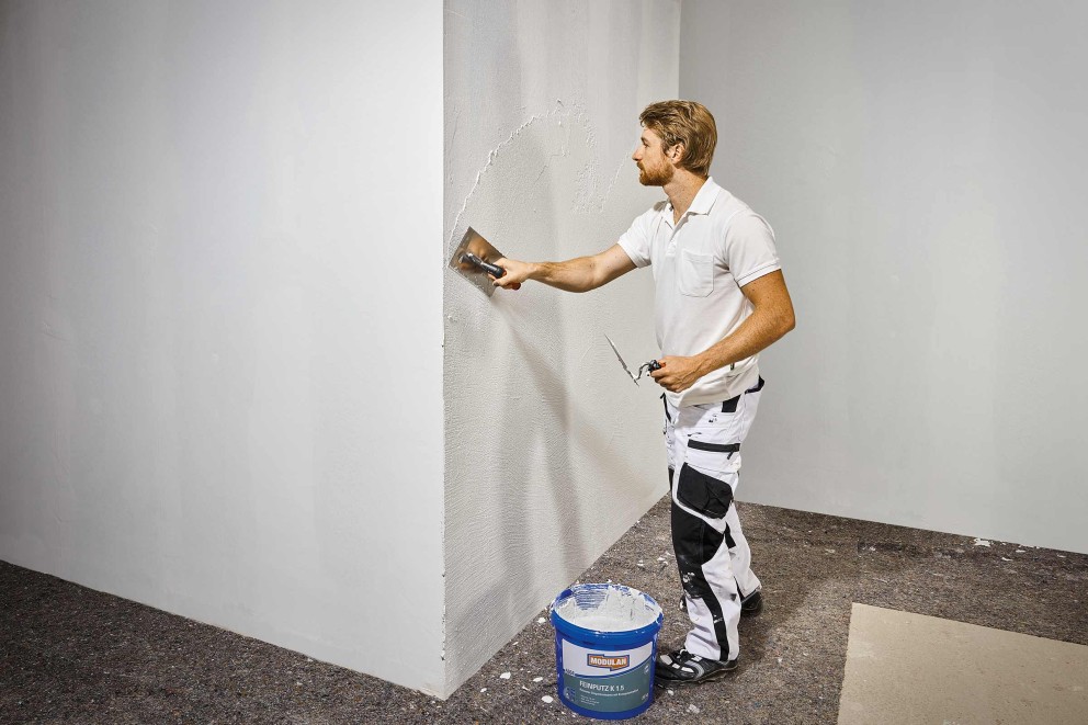 Ein Mann trägt Putz mit einer Glättkelle auf eine Wand auf, ein Eimer Putz steht daneben.