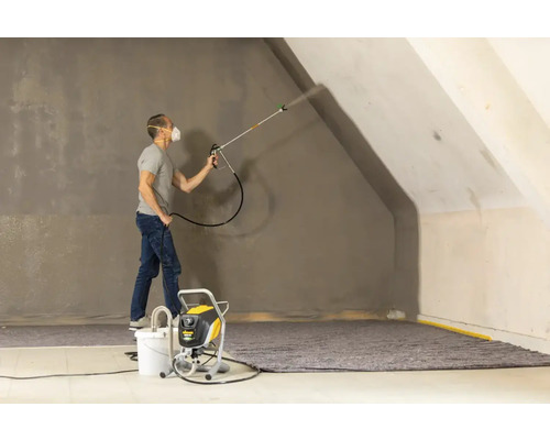 Ein Mann sprüht Farbe mit einem Farbsprühgerät auf eine Wand in einem Raum.