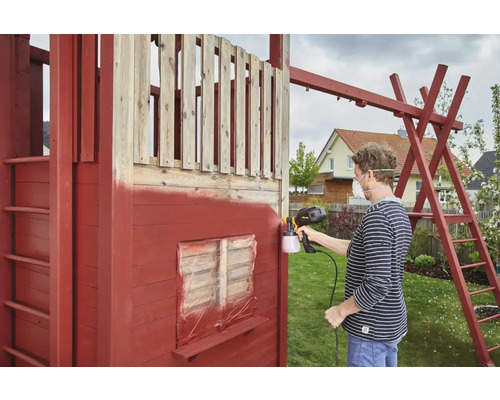 Eine Person sprüht ein hölzernes Spielhaus mit einer Farbspritzpistole an