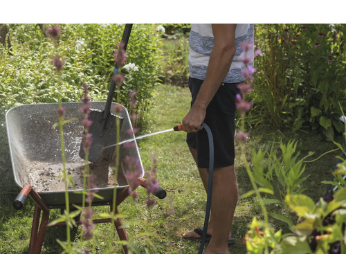 Eine Person reinigt eine Schubkarre mit einem Wasserschlauch in einem Garten, in dem sich auch ein Spaten befindet.