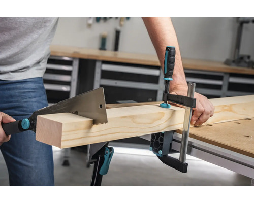 Eine Person sägt ein Holzbrett, das mit einer Schraubzwinge auf einem Werktisch befestigt ist.