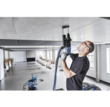 Ein Handwerker bohrt mit einem Bohrhammer ein Loch in die Decke. Ein Staubsauger ist angeschlossen, um Staub zu entfernen. Im Hintergrund befindet sich ein großer Konferenzraum.