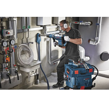 Ein Mann bohrt mit einem Bohrhammer in eine Wand in einem Heizungsraum. Eine Werkzeugkiste und ein Industriesauger stehen bereit.