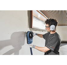 Ein Mann arbeitet mit einem Bohrstaubabsaugungsgerät an einer Wand, wobei er einen Gehörschutz und eine Schutzbrille trägt.