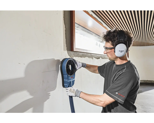 Ein Mann arbeitet mit einem Bohrstaubabsaugungsgerät an einer Wand, wobei er einen Gehörschutz und eine Schutzbrille trägt.