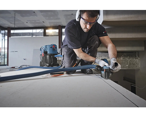 Handwerker schneidet eine Gipsplatte mit einer Mauernutfräse, verbunden mit einem Industriestaubsauger