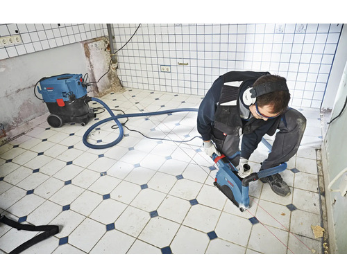Handwerker arbeitet mit einer Mauernutfräse und Staubsauger in einem Raum mit Fliesenboden.