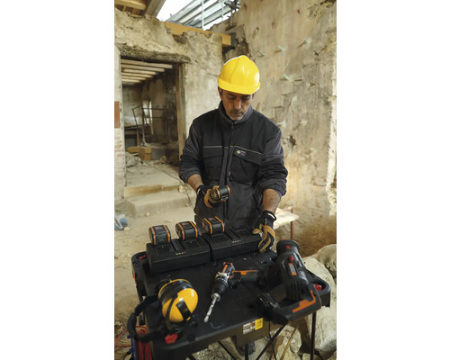 Handwerker mit gelbem Schutzhelm an einem Tisch mit Akkus, Ladegeräten und Werkzeugen in einem Gebäude.