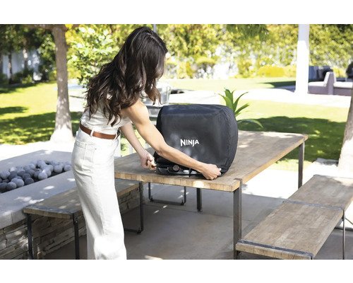 Eine Frau befestigt eine Schutzhülle mit dem Ninja Logo an einem Grill auf einem Tisch im Freien.