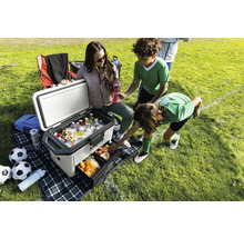 Kühlbox mit Getränken und Snacks auf einer Picknickdecke mit Fußballmotiven, umgeben von Personen auf einem Fußballfeld.