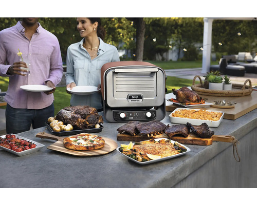 Szene mit einem Ninja Woodfire Outdoor-Ofen und verschiedenen zubereiteten Speisen im Freien.