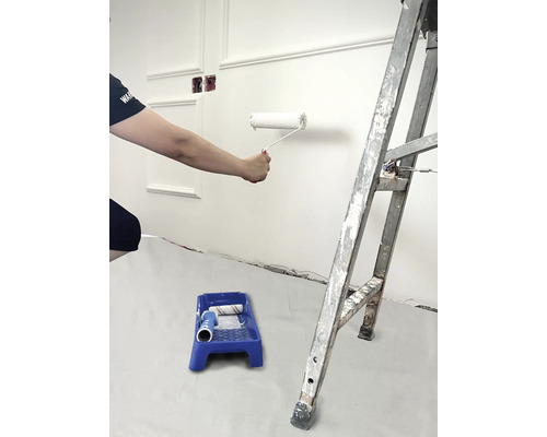 Person streicht Wand mit Farbroller, Farbschale und Leiter im Raum.