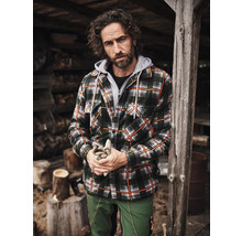 Mann in gefütterter Holzfällerjacke mit Kapuze, Forstjacke und Arbeitshose vor Holzstapel