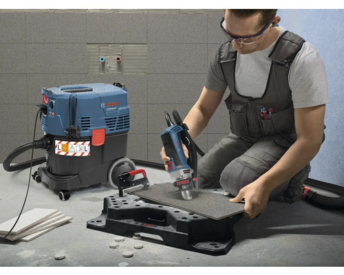 Ein Mann bearbeitet eine Fliese mit einem Fliesenbohrer, während ein Industriesauger bereitsteht, um Staub abzusaugen.