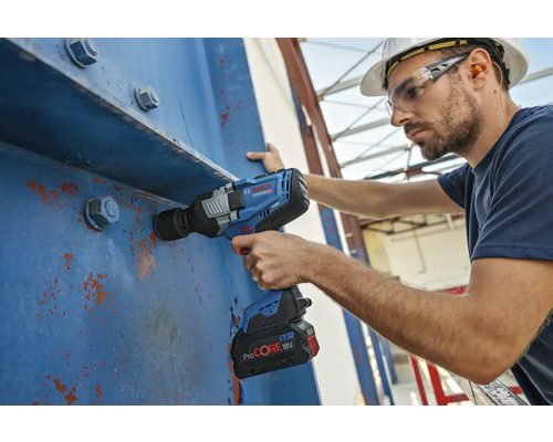 Ein Mann mit Schutzhelm verwendet einen Akku-Schlagschrauber an einer blauen Stahlkonstruktion.