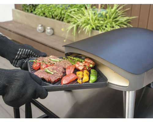 Eine Person mit Grillhandschuhen schiebt eine Grillplatte mit Steak und Gemüse in einen Pizzaofen.