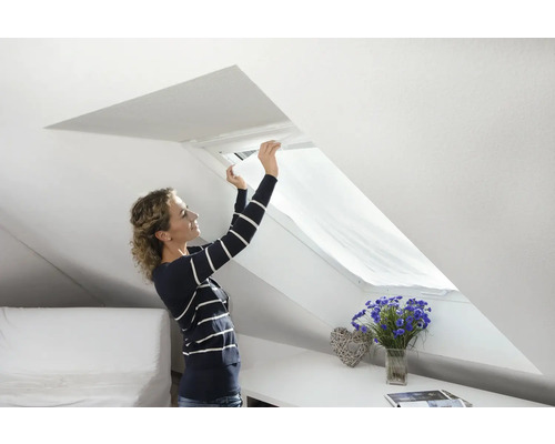 Frau montiert Sonnenschutzrollo an einem Dachfenster in einem hellen Dachzimmer.