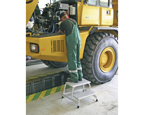 Eine Person steht auf einer zweistufigen Aluminiumleiter und führt Reparaturen an einem gelben Muldenkipper durch.