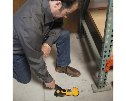 Ein Mann kniet und verwendet einen gelben Metallsucher auf einem Betonboden.