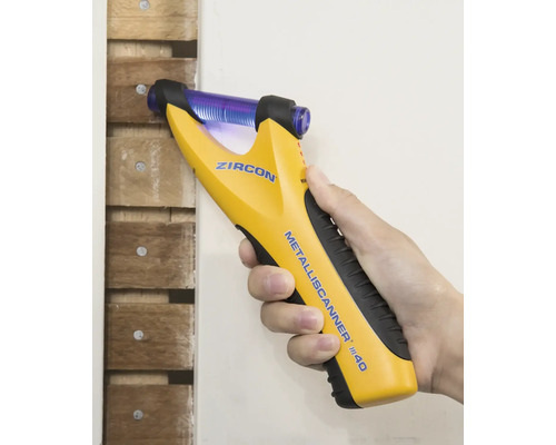 Eine Hand hält einen gelben Metallscanner an einer Holzwand, um Metall zu erkennen.