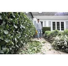 Ein Mann benutzt einen Laubbläser, um einen gepflasterten Weg von Blättern zu befreien, neben einer geschnittenen Hecke und einem Haus.