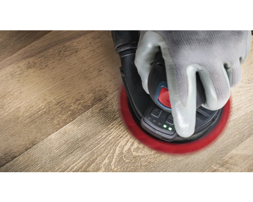 Eine Hand mit Handschuh bedient einen Exzenterschleifer auf einem Holzboden.