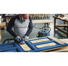 Eine Person schleift mit einem Exzenterschleifer eine blaue Holztür, unterstützt von einem Staubsauger.