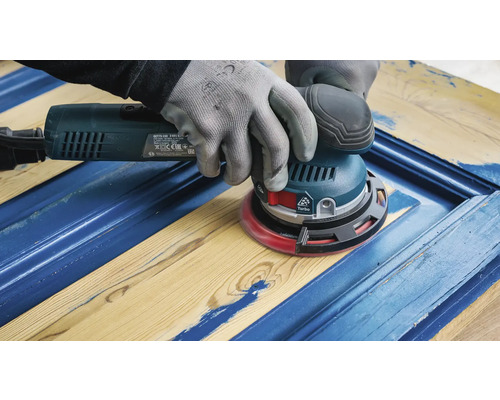 Ein Handwerker schleift mit einem Exzenterschleifer eine blaue Holztür.