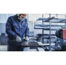 Ein Mann schleift ein Metallstück mit einer Schleifmaschine in einer Werkstatt. Er trägt Schutzkleidung wie Gehörschutz, Schutzbrille, eine Maske und Handschuhe.