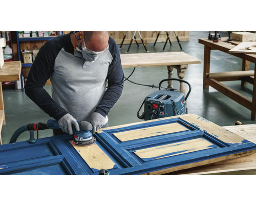 Eine Person schleift mit einem Exzenterschleifer eine blaue Holztür in einer Werkstatt.