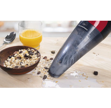 Ein Cleanmaxx Handstaubsauger saugt verschüttetes Müsli mit Milch auf einem Holztisch auf. Im Hintergrund sind eine Schüssel Müsli und ein Glas Orangensaft zu sehen.