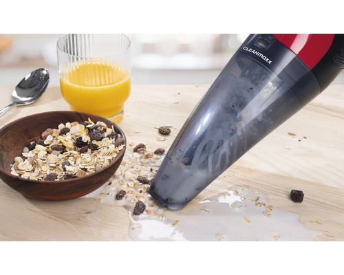 Ein Cleanmaxx Handstaubsauger saugt verschüttetes Müsli mit Milch auf einem Holztisch auf. Im Hintergrund sind eine Schüssel Müsli und ein Glas Orangensaft zu sehen.