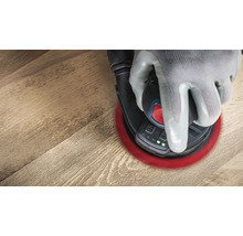 Eine Hand mit Schutzhandschuh bedient einen Exzenterschleifer auf einer Holzoberfläche.