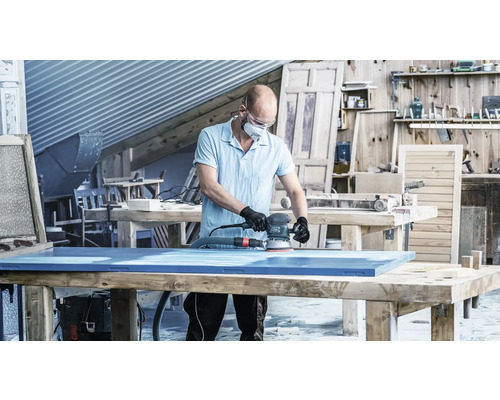 Ein Mann schleift eine blaue Holzplatte mit einem Exzenterschleifer in einer Werkstatt.