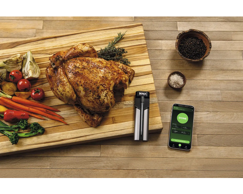 Szene mit gebratenem Hähnchen, Gemüse, Fleischthermometer und Smartphone auf einem Schneidebrett