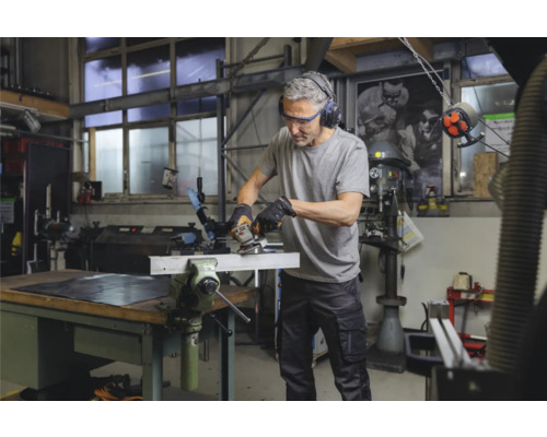 Ein Mann arbeitet in einer Werkstatt mit einem Winkelschleifer an einem Metallstück, das in einem Schraubstock eingespannt ist. Er trägt dabei Sicherheitsbrille und Gehörschutz.