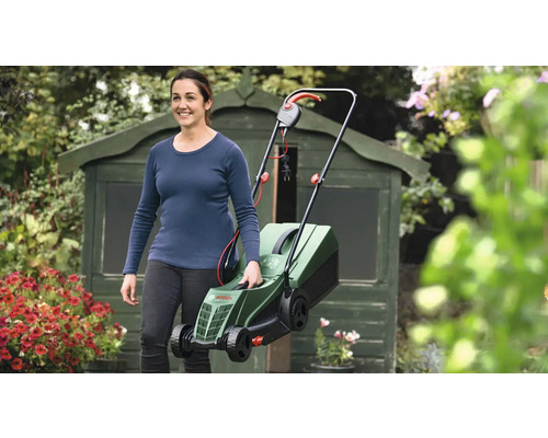 Frau trägt einen elektrischen Rasenmäher im Garten vor einem Geräteschuppen.