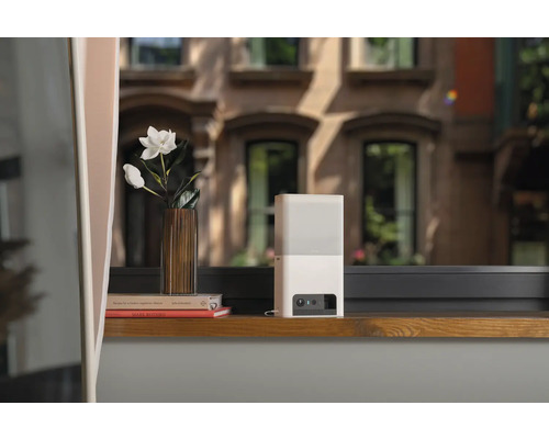 Weiße Sicherheitskamera auf einer Fensterbank mit Blumen und Büchern im Hintergrund.