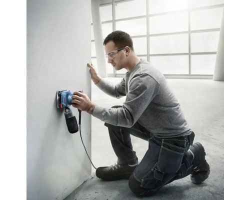 Ein Mann schleift eine Wand mit einem Elektroschleifer in einem hellen Raum.