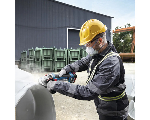 Ein Mann mit Schutzhelm, Schutzbrille und Atemschutzmaske schleift mit einem Akku-Winkelschleifer ein Werkstück.