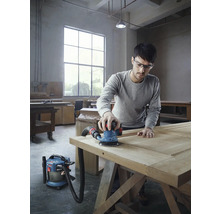 Ein Mann schleift eine Holztür mit einem Exzenterschleifer und einem Staubsauger in einer Werkstatt.