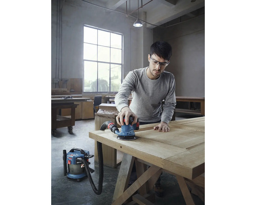 Ein Mann schleift eine Holztür mit einem Exzenterschleifer und einem Staubsauger in einer Werkstatt.