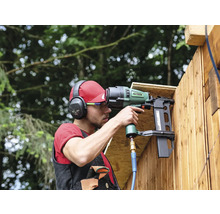 Ein Mann befestigt Holzlatten mit einem Druckluftnagler an einer Wand. Er trägt dabei Schutzkleidung wie Kopfhörer und Schutzbrille.
