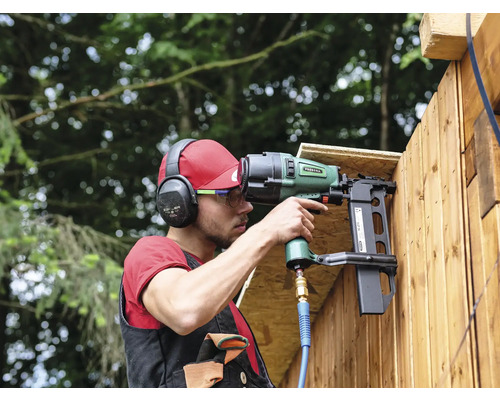Ein Mann befestigt Holzlatten mit einem Druckluftnagler an einer Wand. Er trägt dabei Schutzkleidung wie Kopfhörer und Schutzbrille.