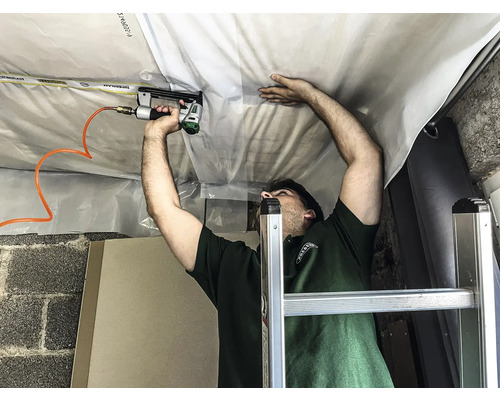 Ein Mann befestigt eine Folie mit einem Druckluftnagler an einer Decke, stehend auf einer Leiter.