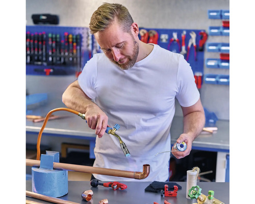 Ein Mann lötet Kupferrohre mit einem Lötkolben in einer Werkstatt, Kupferfittings und Werkzeuge liegen auf dem Tisch.